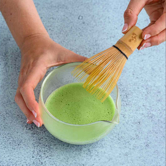 Person whisking green matcha powder in a glass bowl with a wooden whisk on a light blue surface.