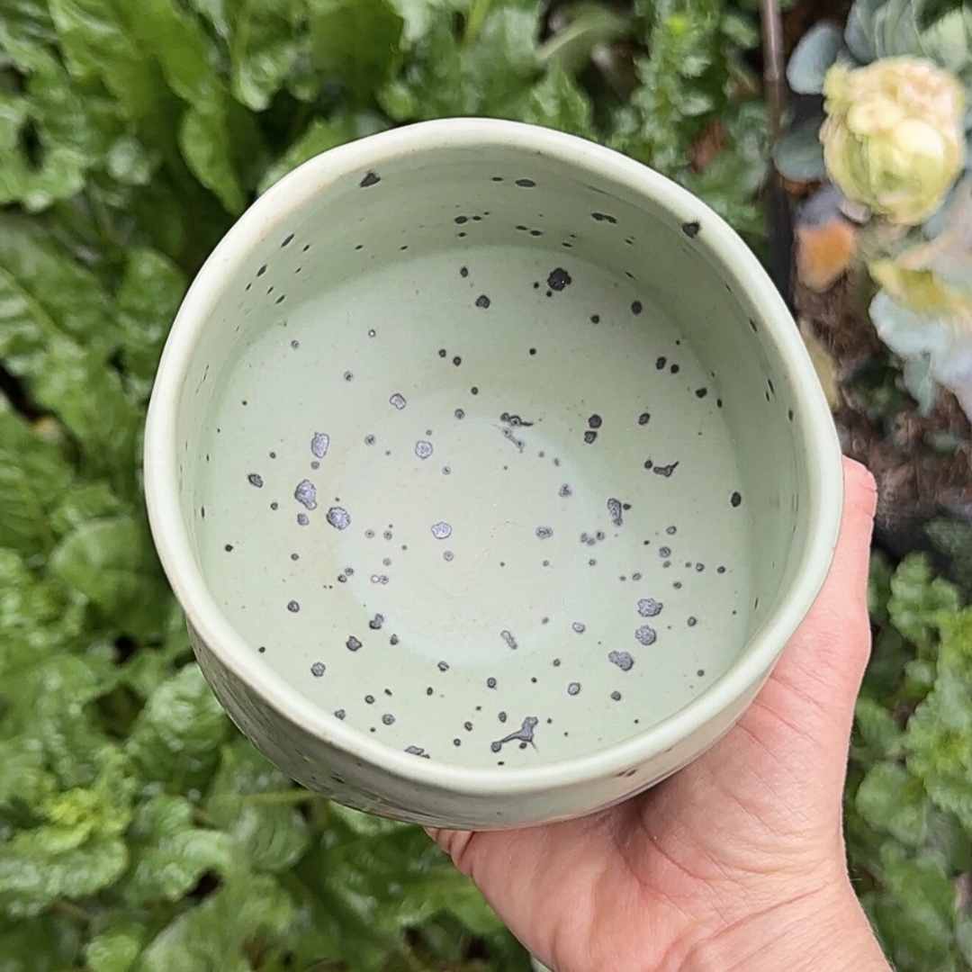 Hand holding a green speckled ceramic bowl against a natural background