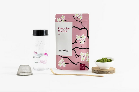 A pink stand-up pouch of Everyday Matcha from the brand Matcha Yu Tea displayed on a white background alongside a clear glass jar with a black lid, a metal strainer, a wooden spoon, and a small white bowl of green matcha on a wooden coaster.