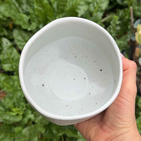 Hand holding a white ceramic bowl with speckled texture against a green leafy background
