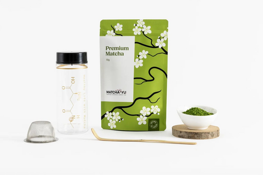 A package of matcha tea with a green pouch decorated with white cherry blossoms, shown alongside a clear bottle with a black cap, a wooden spoon, and a small bowl of green matcha powder on a white background.