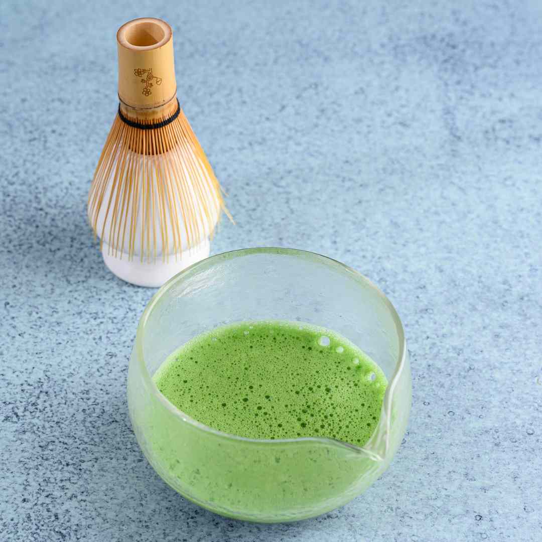 Bowl of green matcha tea with a bamboo whisk on a light blue textured surface