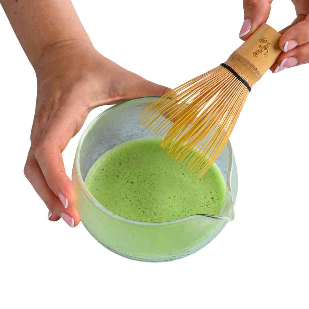 Person using a wooden whisk to stir green matcha tea in a glass bowl.