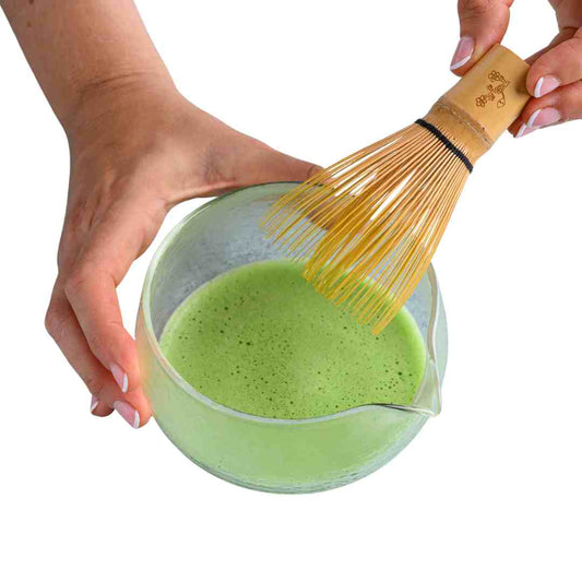 Person using a wooden whisk to stir green matcha tea in a glass bowl.