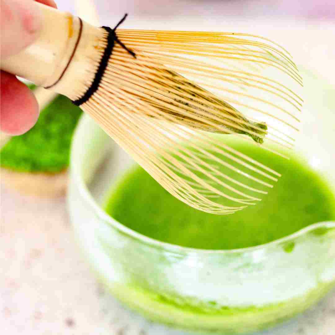 Wooden whisk being used to stir green tea in a glass bowl.