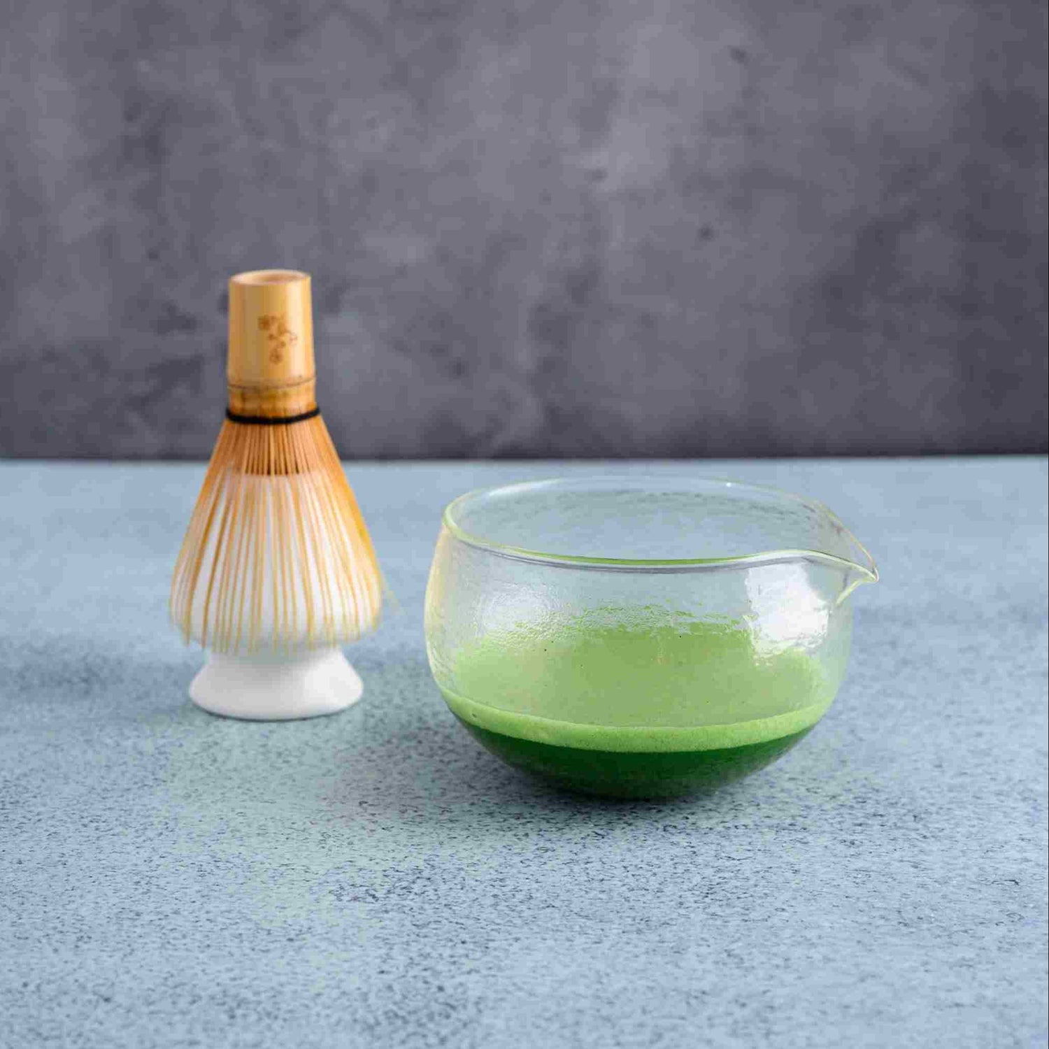 Bamboo whisk and glass container with Matcha liquid on a gray surface