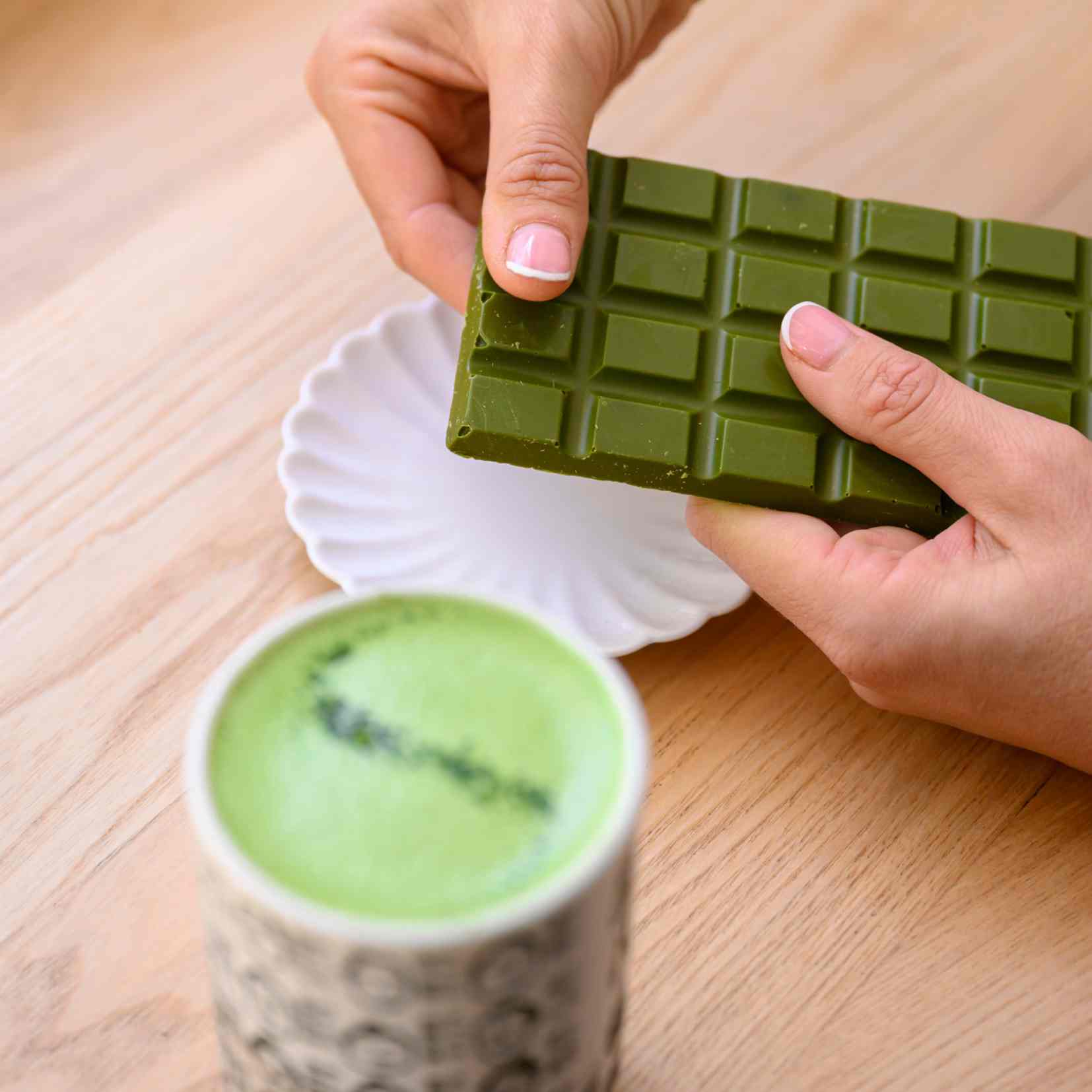 Green matcha chocolate bar held by a hand next to a cup of matcha on a wooden surface