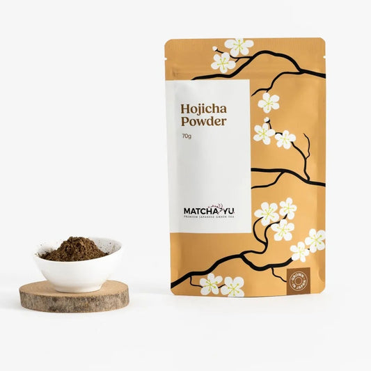 A stand-up pouch of Matcha·yu Hojicha Powder with a tan and white design featuring cherry blossom branches, shown next to a small white bowl filled with dark green powder on a wooden coaster against a white background.