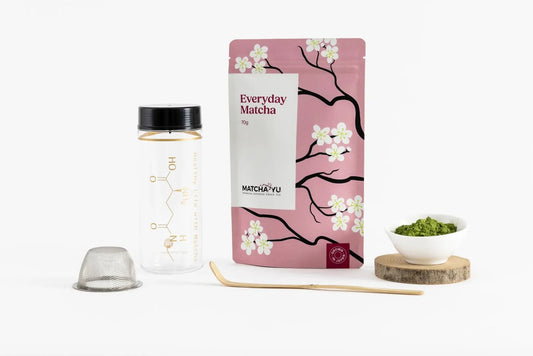 A matcha tea kit on a white background, featuring a pink stand-up pouch with cherry blossom illustrations labeled 'Everyday Matcha' a clear bottle with a black cap, a metal tamper, a wooden spoon, and a small white bowl of green matcha powder on a wooden coaster.