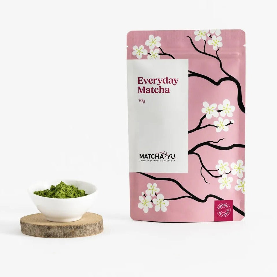 A pink stand-up pouch of Matcha. Everyday Matcha with cherry blossom illustrations, displayed next to a small white bowl of green matcha tea on a wooden coaster against a white background.