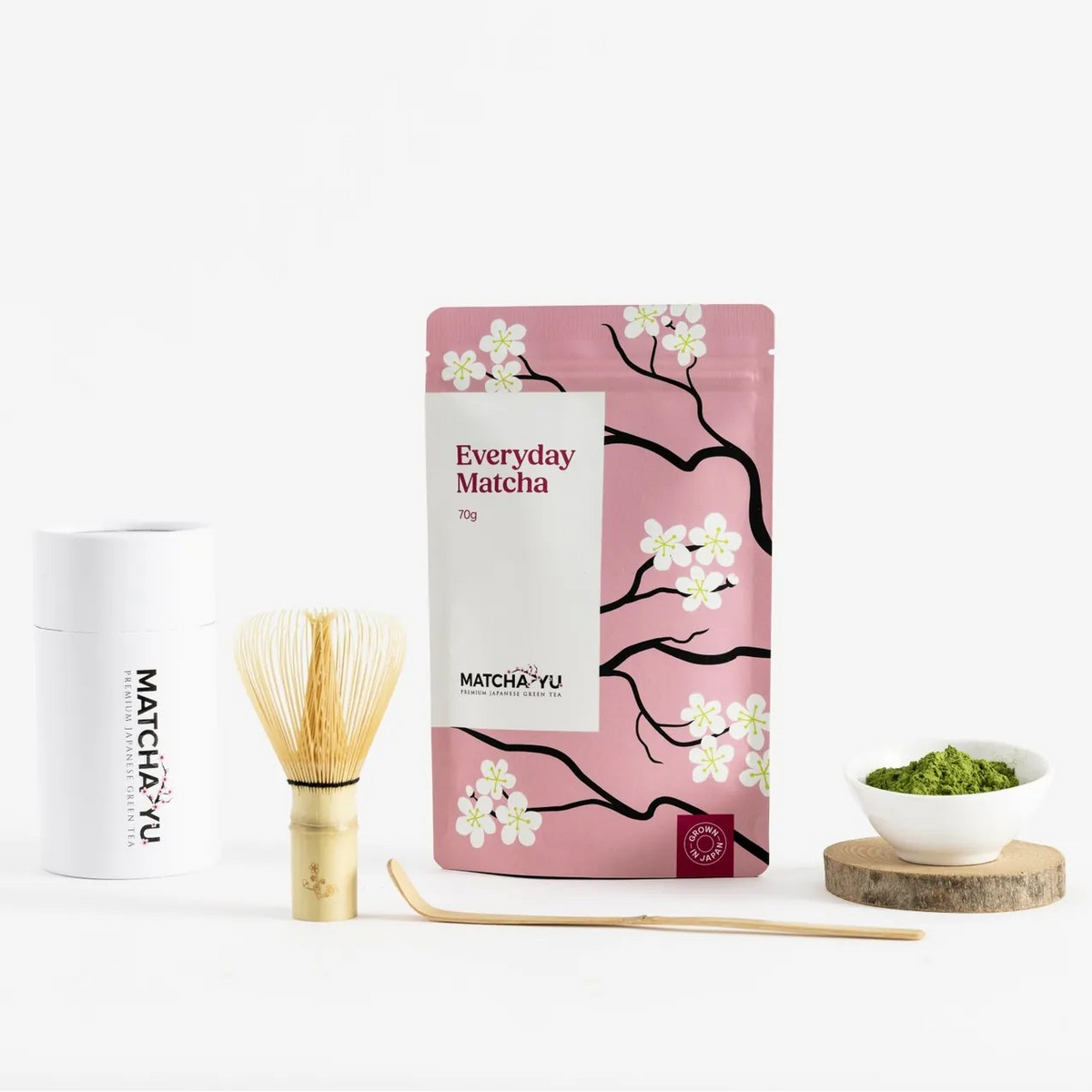 A matcha tea product set on a clean white background, featuring a pink stand-up pouch with cherry blossom illustrations labeled 'Everyday Matcha' and 'Matcha Yu Tea', a white cylindrical tin, a bamboo whisk, a wooden scoop, and a small bowl of green matcha on a wooden coaster.