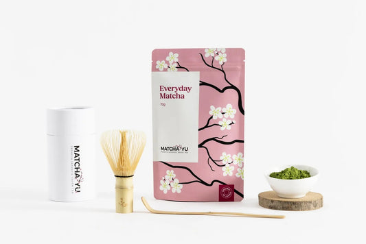 A matcha tea product set on a clean white background, featuring a pink stand-up pouch with cherry blossom illustrations labeled 'Everyday Matcha' and 'matcha yu tea', a white cylindrical tin, a bamboo whisk, a wooden scoop, and a small bowl of green matcha on a wooden coaster.