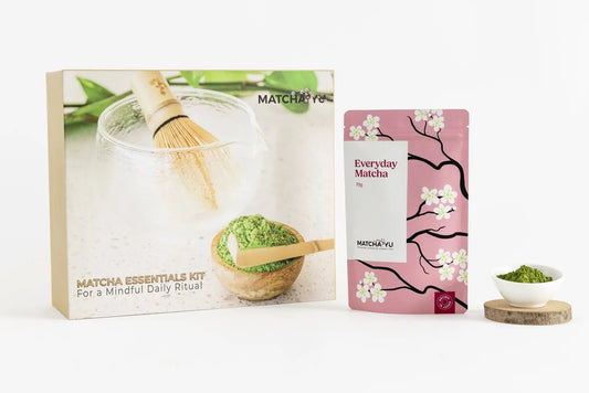 Matcha Essentials Kit by Matcha Yu Tea featuring a cardboard box with a whisk and green matcha illustration, a pink stand-up pouch with cherry blossom graphics labeled 'Everyday Matcha', and a small bowl of green matcha powder on a wooden coaster, all on a clean white background.