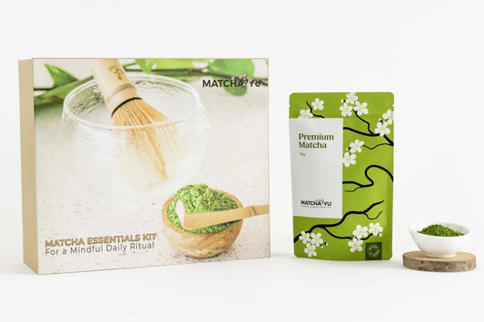 A matcha tea gift set on a clean white background, featuring a cardboard box labeled 'Matcha Essentials Kit', a green stand-up pouch marked 'Premium Matcha' with white cherry blossoms, a bamboo whisk, and a small bowl of matcha powder on a wooden coaster.
