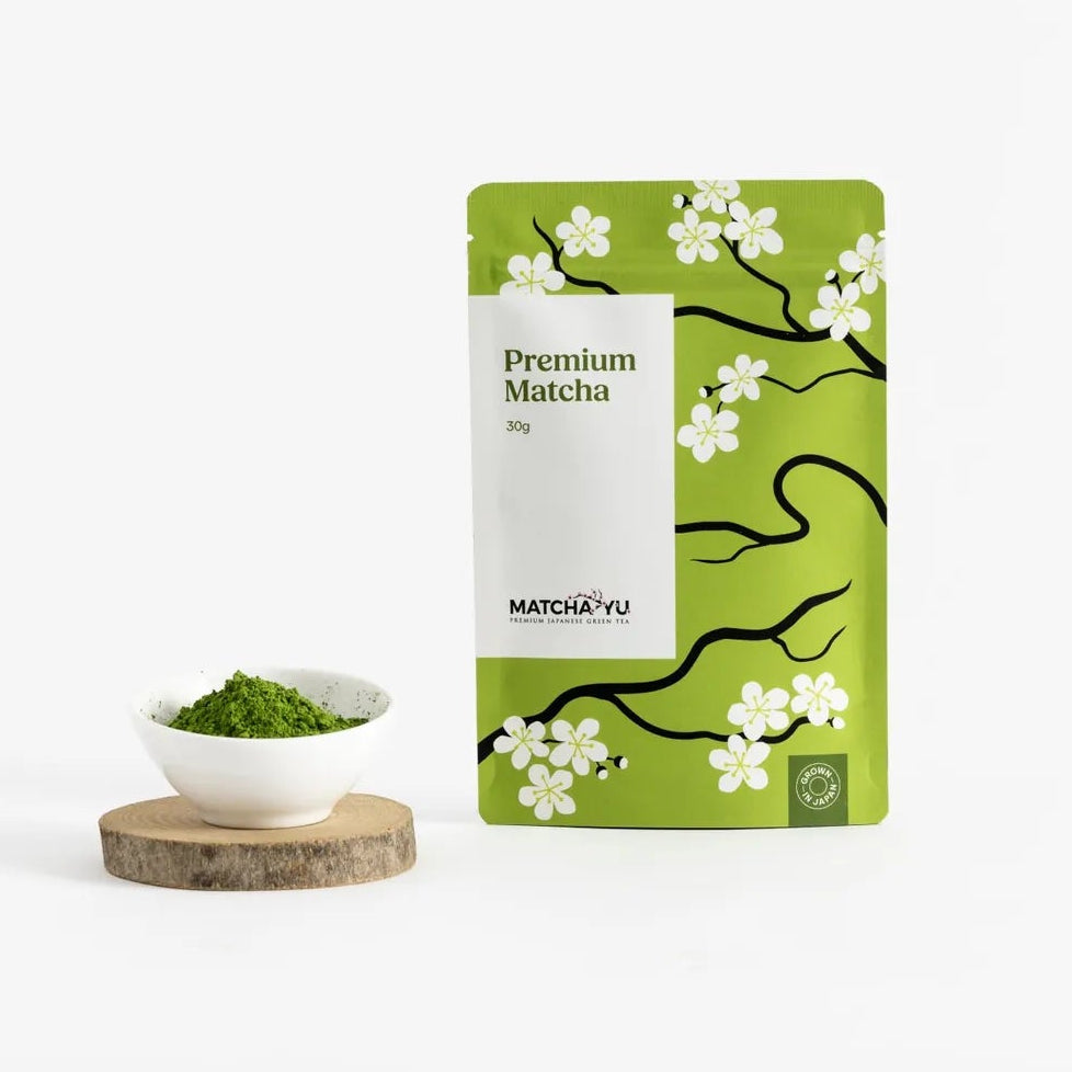 A green stand-up pouch of Matcha yu Tea Premium Matcha with white cherry blossom illustrations, displayed next to a small white bowl of green matcha powder on a wooden coaster against a clean white background.