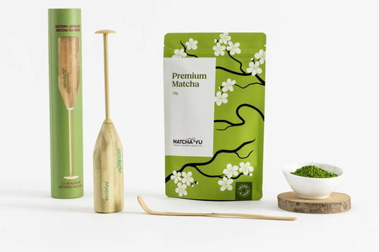 A matcha tea set arrangement on a white background, including a green cylindrical box, a bamboo whisk, a green pouch labeled 'Premium Matcha' with white cherry blossom illustrations, a bamboo scoop, and a small white bowl of green matcha powder on a wooden coaster.
