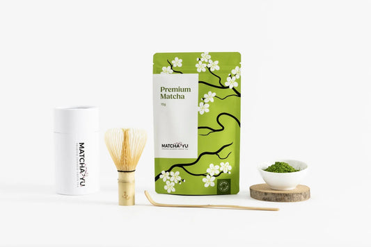 A matcha tea set on a clean white background, featuring a green and white pouch of Matcha Yu Tea Premium Matcha, a white cylindrical tin, a bamboo whisk, a bamboo scoop, and a small white bowl of green matcha powder on a wooden coaster.