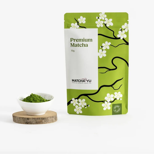 A stand-up pouch of Matcha Yu Tea Premium Matcha tea stands on a white surface against a white background, next to a small white bowl filled with green matcha powder on a round wooden coaster.